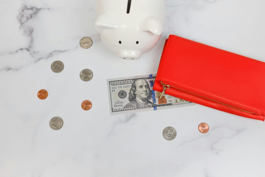A white porcelain piggy bank on a marble table with coins, a paper bill, and a red wallet.
