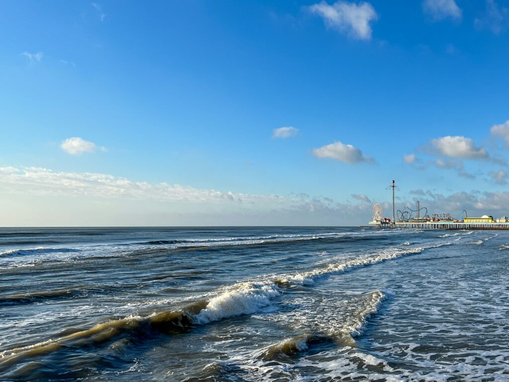 Bright blue sky sunny day over the blue ocean shoreline in the morning along Galveston Beach, Texas with Pleasure Pier in the distance.